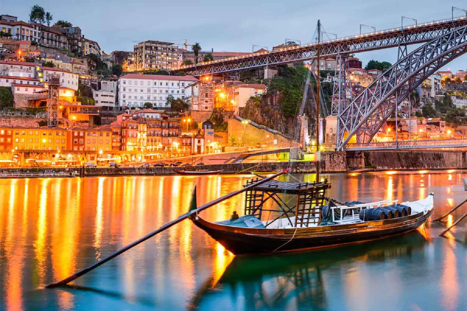 Traditional wooden boat moored in a river with illuminated buildings and a bridge in the background during twilight.